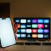 Streaming platforms home entertainment smartphone blank screen mockup television. Hand holding modern mobile device empty display foreground large smart television interface background living room interior technology lifestyle media consumption editorial concept