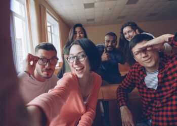 Point of view shot of funny attractive young men and women taking selfie in lecture hall, posing with textbook and showing hand gestures and expressive faces.