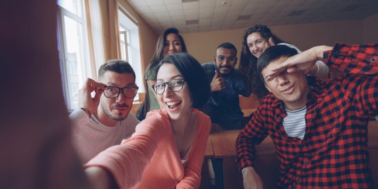 Point of view shot of funny attractive young men and women taking selfie in lecture hall, posing with textbook and showing hand gestures and expressive faces.