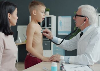 Pediatrician senior man in white gown is examining child in hospital using stethoscope while mother attractive young woman is watching boy in office.